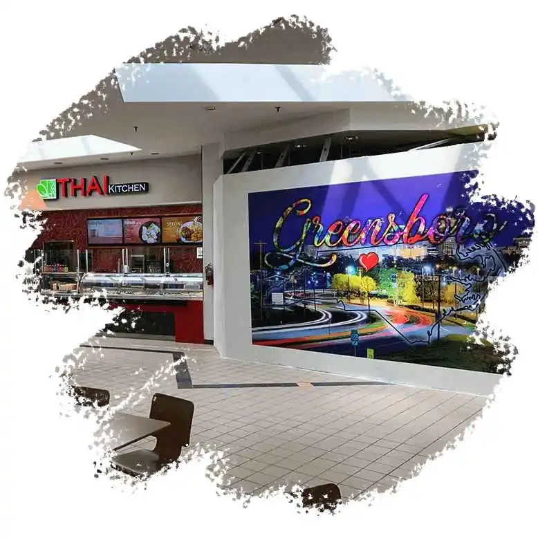 Inside a shopping mall, a Thai Kitchen eatery is visible beside a colorful wall mural showcasing "Greensboro" with a heart symbol.