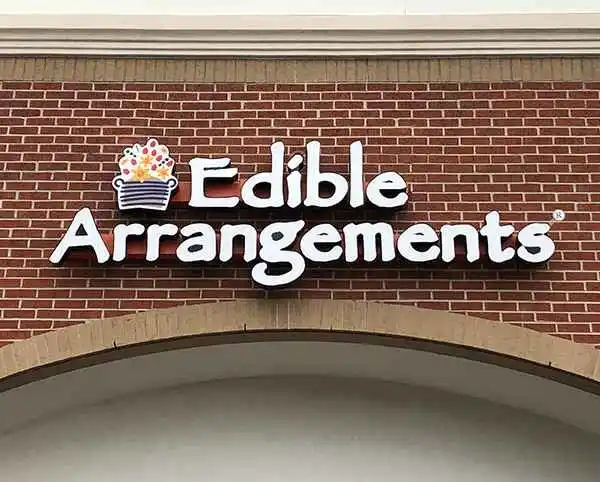 Sign for Edible Arrangements displayed on a brick wall, featuring a colorful fruit bouquet logo above the brand name in white letters.